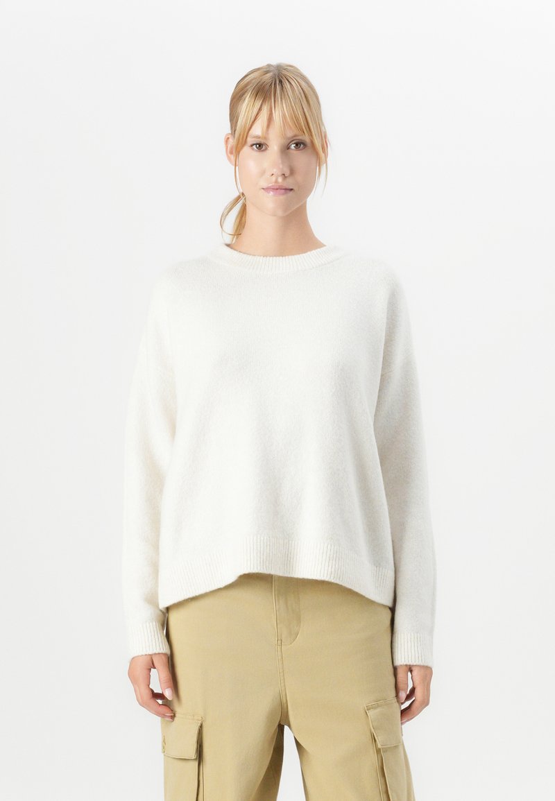 Giovane donna con capelli biondi raccolti in una coda di cavallo, che indossa un maglione bianco a maniche lunghe e pantaloni cargo beige, in piedi davanti a uno sfondo bianco semplice.