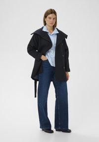 Tmavomodrý vlnený kabát s veľkým golierom, dvojradovými gombíkmi a opaskom, doplnený svetlomodrou košeľou a širokými džínsami s voľnými nohavicami.