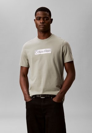 Hombre de pie con las manos en los bolsillos vistiendo una camiseta Calvin Klein gris claro y pantalones negros sobre un fondo gris liso.