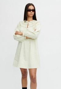 Vrouw met lang, donker haar, die een zonnebril draagt en een losse crèmekleurige jurk met een gegatherde yoke en lange mouwen aan heeft, staand met de armen gekruist.