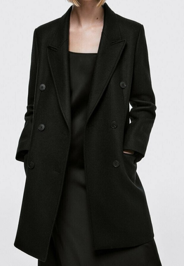 Femme portant un manteau en laine noir à double boutonnage avec un col cranté sur une robe noire, mains dans les poches du manteau.