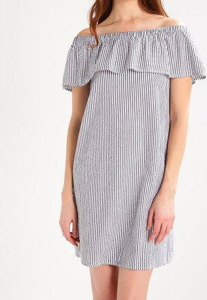 Kvinde iført en kort off-shoulder kjole med lodrette sorte og hvide striber og flæsekant ved halsudskæringen.