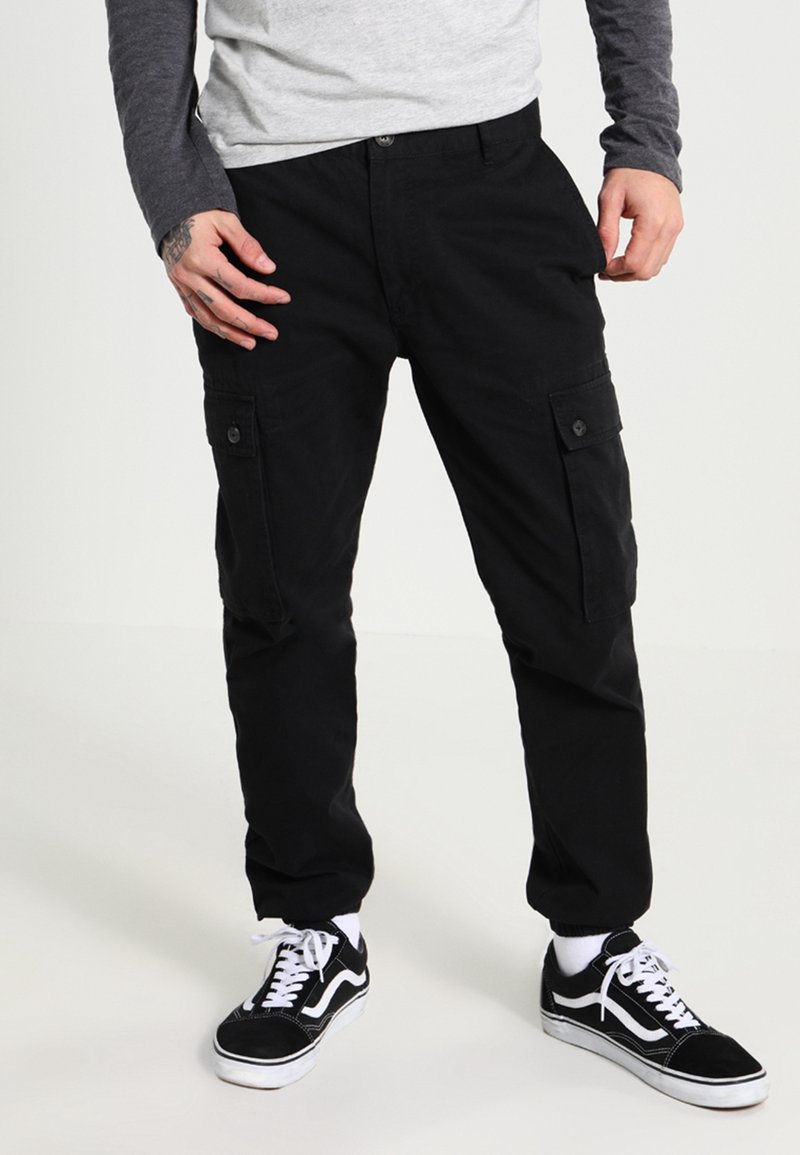 Homme portant un pantalon cargo noir avec poches latérales boutonnées et des baskets basses noires avec lacets blancs et semelles blanches, debout sur un fond blanc.
