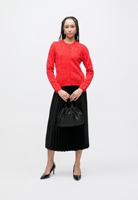 Rode gebreide cardigan met bloemmotief, dichtgeknoopt met knopen, gecombineerd met een zwarte geplooide rok en een zwarte leren tas. Zwarte hakken maken de outfit compleet.