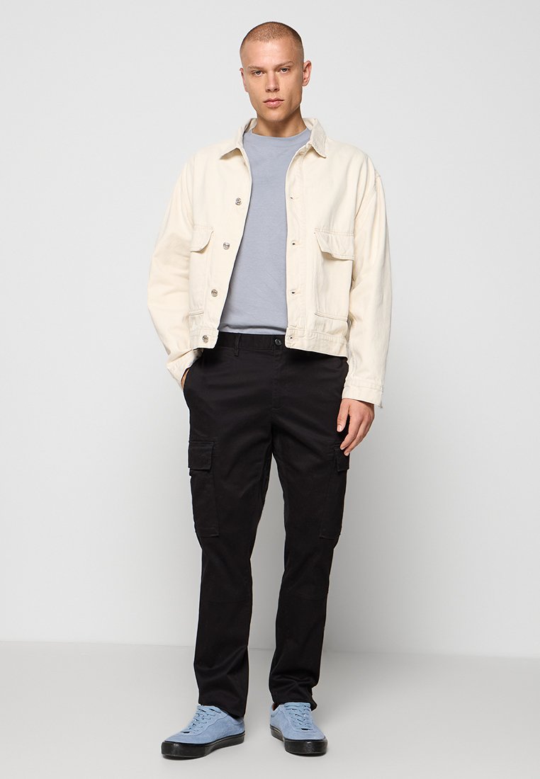 Veste en denim crème clair avec deux poches avant, t-shirt gris, pantalon cargo noir et baskets bleues sur fond uni.