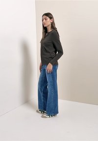 Pitkähihainen tumma paita yhdistettynä leveälahkeisiin sinisiin farkkuihin. Asussa on vaaleanharmaat sandaalit, joissa on vihreät raidat sukissa. Yksivärinen tausta.