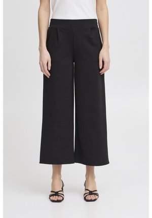 Mujer con pantalones negros cortos de cintura alta y piernas anchas, camiseta blanca sin mangas y sandalias negras con dedos abiertos y tiras finas.