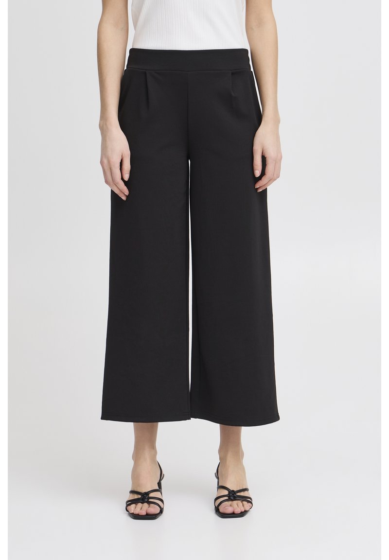 Femme portant un pantalon noir taille haute, jambes larges et coupé, un top blanc sans manches et des sandales noires à bouts ouverts avec fines brides.