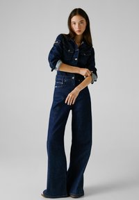 Veste en denim foncé avec un jean large assorti. La veste possède une fermeture à boutons et des détails en patch. Texture lisse, coupe classique et tons bleus.