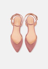 Zapatos planos de punta afilada en color rosa claro, con una correa en el tobillo, fabricados en un material similar al ante, que presenta una textura suave y un diseño minimalista.