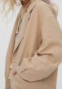 Chaqueta beige, texturizada, con un corte holgado, grandes solapas y bolsillos laterales. Superpuesta sobre una sudadera pastel, mostrando un tejido suave y un diseño informal.