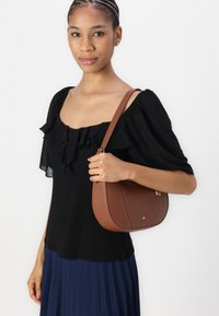 Braune Lederhandtasche mit einer geschwungenen Form, ausgestattet mit einem einfachen Verschluss und einem Tragegurt. Das Model trägt ein schwarzes Oberteil mit Rüschensäumen und einen marineblauen Faltenrock.