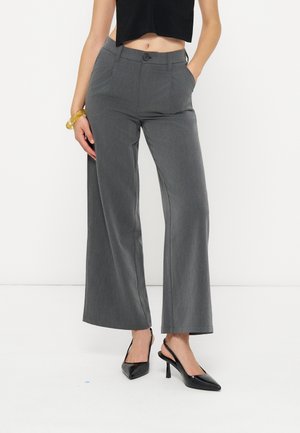 Kvinde iført grå bukser med vide ben, sort cropped top, sorte slingback hæle med spids tå og et gult armbånd, stående med den ene hånd i lommen.