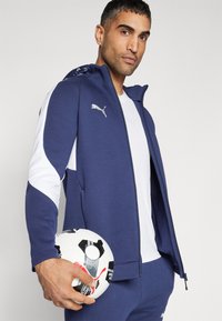 Tengerészkék sportdzseki fehér részletekkel, kapucnival és zippzárral. A modellen egy fekete-fehér focilabda van.