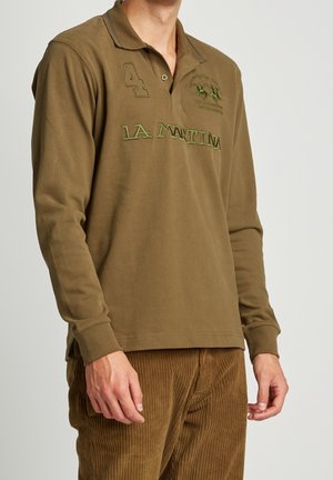 Homme portant un polo à manches longues vert olive avec le texte "LA MARTINA" et le numéro 4, associé à un pantalon en velours côtelé marron.