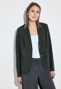 Blazer verde scuro con un design su misura, due tasche frontali e chiusure con bottoni. Indossato sopra una maglietta bianca e abbinato a pantaloni grigi.