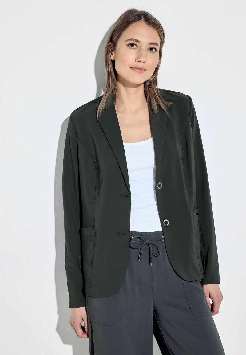 Blazer verde scuro con un design su misura, due tasche frontali e chiusure con bottoni. Indossato sopra una maglietta bianca e abbinato a pantaloni grigi.