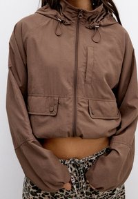 Brown cropped jacket with a hood, zip closure, and two front pockets. Made of lightweight material, features adjustable drawstrings.