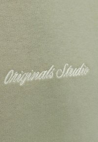 Svetlo zelena pletena tkanina z belo vezenim kurzivnim besedilom, na katerem piše "Originals Studio."