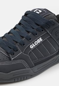 Black sneaker with textile and synthetic upper, textured accents, white stitching, and logo. Features dark laces and a padded tongue.