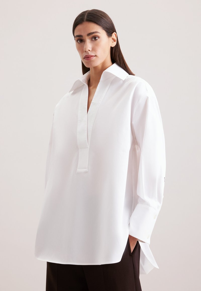 Camicia bianca a maniche lunghe con colletto e dettaglio della bottoniera. Presenta una vestibilità ampia, una texture liscia e spacchi laterali all'orlo.