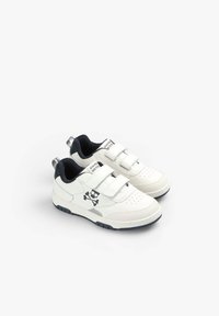 Weiße Sneakers mit Klettverschlüssen, schwarzem Innenfutter, durchlässiger Zehenpartie und einem Totenkopf-und-Kreuzbein-Druck an der Seite. Robuste Gummisohle.