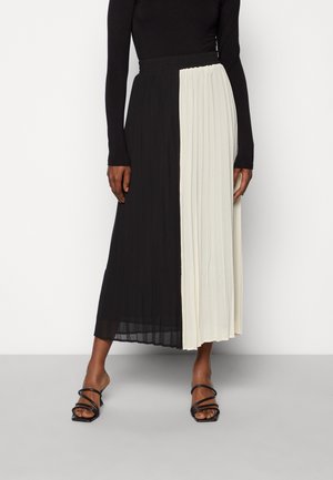 Nainen, jolla on päällään musta pitkähihainen paita ja mustavalkoinen pliseerattu midihame, yhdistettynä mustiin remmikenkiin.