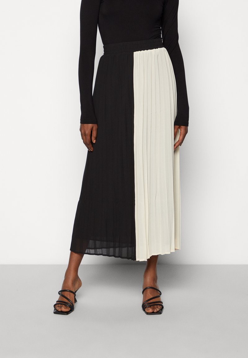 Femme portant un haut noir à manches longues et une jupe midi plissée noire et blanche, associée à des talons noirs à brides.