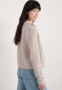 Vaaleanbeige neulepusero, jossa on erilaisia kuvioita, resorit hihansuissa ja kauluksessa. Yhdistetty vaaleansinihin farkkuihin. Takaa nähtynä.