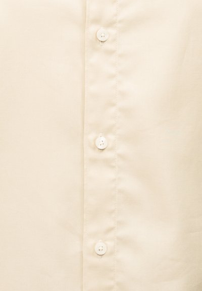 Κοντινό πλάνο μπεζ υφάσματος με τρία λευκά κουμπιά τοποθετημένα κάθετα σε μία λωρίδα πουκάμισου.