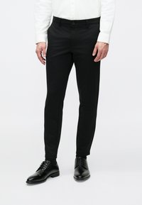 Schwarze, maßgeschneiderte Hose aus glattem Stoff, mit Knopfverschluss, schmaler Passform und kombiniert mit schwarzen Anzugschuhen.