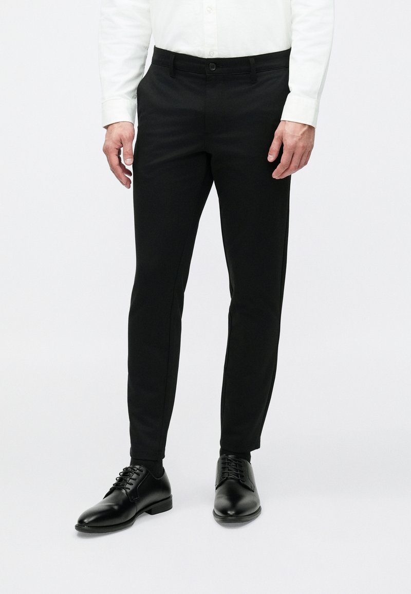 Schwarze, maßgeschneiderte Hose aus glattem Stoff, mit Knopfverschluss, schmaler Passform und kombiniert mit schwarzen Anzugschuhen.