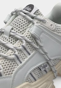 Λευκός αθλητικός παπούτσι από δίχτυ και δέρμα με κλείσιμο με κορδόνια και ετικέτες με λογότυπο στη γλώσσα και το πλάι, με υφή μπεζ σόλας και αντανακλαστικές ρίγες.