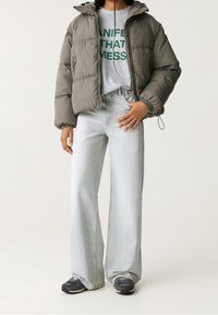 Pufová bunda v olivově zelené, šedé tričko s nápisem, světlé modré kalhoty s širokými nohavicemi a černé tenisky s bílou podešví na neutrálním pozadí.