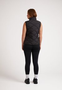 Svart vadderad väst med hög krage, med ett quiltat mönster. Bärs över svarta leggings och svarta sneakers, med vita strumpor synliga.