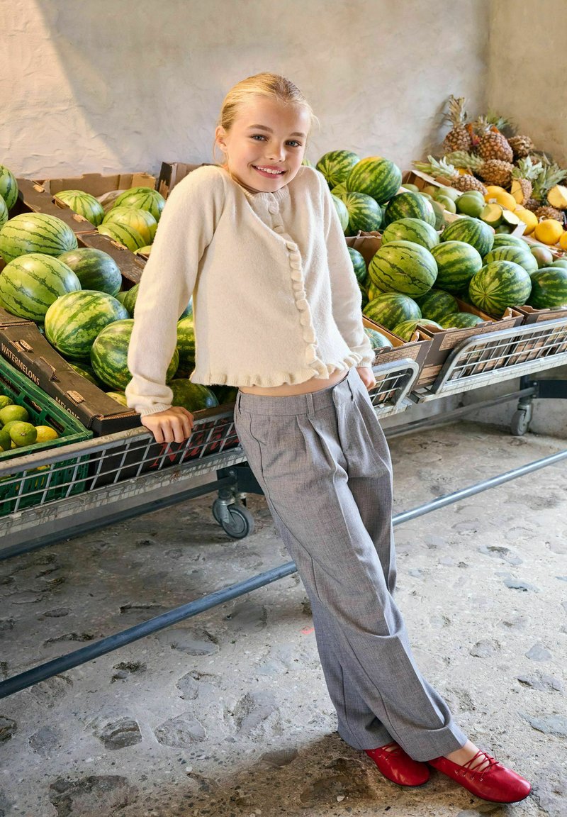 Jeune fille appuyée contre un étalage de pastèques, souriante, portant un pull beige, un pantalon gris et des chaussures rouges, dans un marché de fruits.
