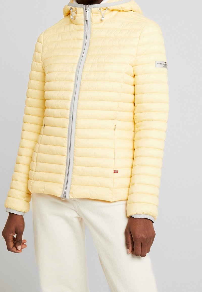 Homme portant une veste matelassée jaune clair avec capuche et fermeture éclair devant, assortie à un pantalon blanc.