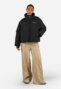 Svart polstret jakke med høy krage og glidelås, med ribbestrikkede mansjetter og en hvit logo på venstre side. Kombinert med beige vide bukser.