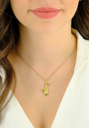 Pendentif saxophone en or sur une chaîne délicate, présentant des détails sophistiqués et une finition lisse, porté sur un cou nu au-dessus d'un chemisier blanc.