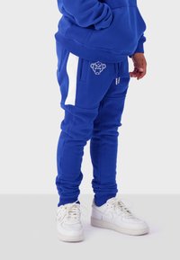 Blue sweatpants with white side stripes, fabric fleece texture, adjustable waist, and a logo on the pocket. Paired with white sneakers.