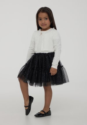 Top bianco a maniche lunghe con dettagli arricciati, gonna in tulle nera con glitter e ballerine in vernice nera. Bambina in piedi che posa con sicurezza.