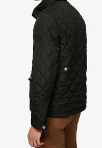 Giacca nera trapuntata con colletto rialzato, polsini con bottoni e tasche laterali. Il materiale è lucido e ha una texture leggermente satinata.