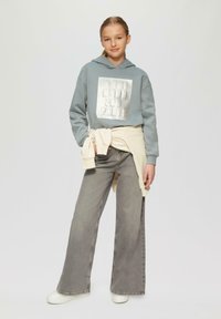 Grijze hoodie met de tekst "IN THE MOOD FOR PARTY", gecombineerd met lichtgrijze wijde jeans. Sweatshirt om de taille geknoopt. Witte sneakers.