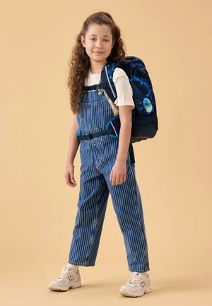 Girl with curly hair wearing striped denim overalls, white sneakers, and carrying a large blue backpack standing on a beige background.