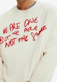 Sudadera blanca con texto bordado en rojo que dice "SOMOS UNO PERO NO SOMOS LO MISMO". Tiene un cuello redondo y mangas largas.