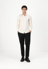 Camasa bej deschis cu nasturi, având un model texturat, asociată cu pantaloni negri cu croi ajustat și pantofi negri, expusă pe un fundal neutru.