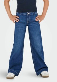 Personne se tenant avec les mains sur les hanches, portant un pantalon en denim large bleu, un haut sans manches noir et des baskets blanches, sur un fond blanc.