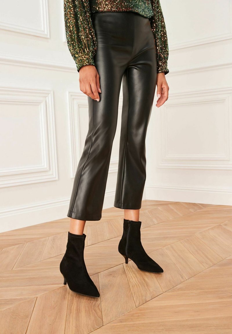 Femme portant un pantalon court en cuir synthétique noir et des bottines noires à bouts pointus sur un sol en bois avec un mur blanc à panneaux.