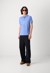 Cămașă polo de culoare albastru deschis cu mâneci scurte, logo mic pe piept, asociată cu pantaloni negri cu croială largă și pantofi slip-on maro. Ochelari de soare incluși.