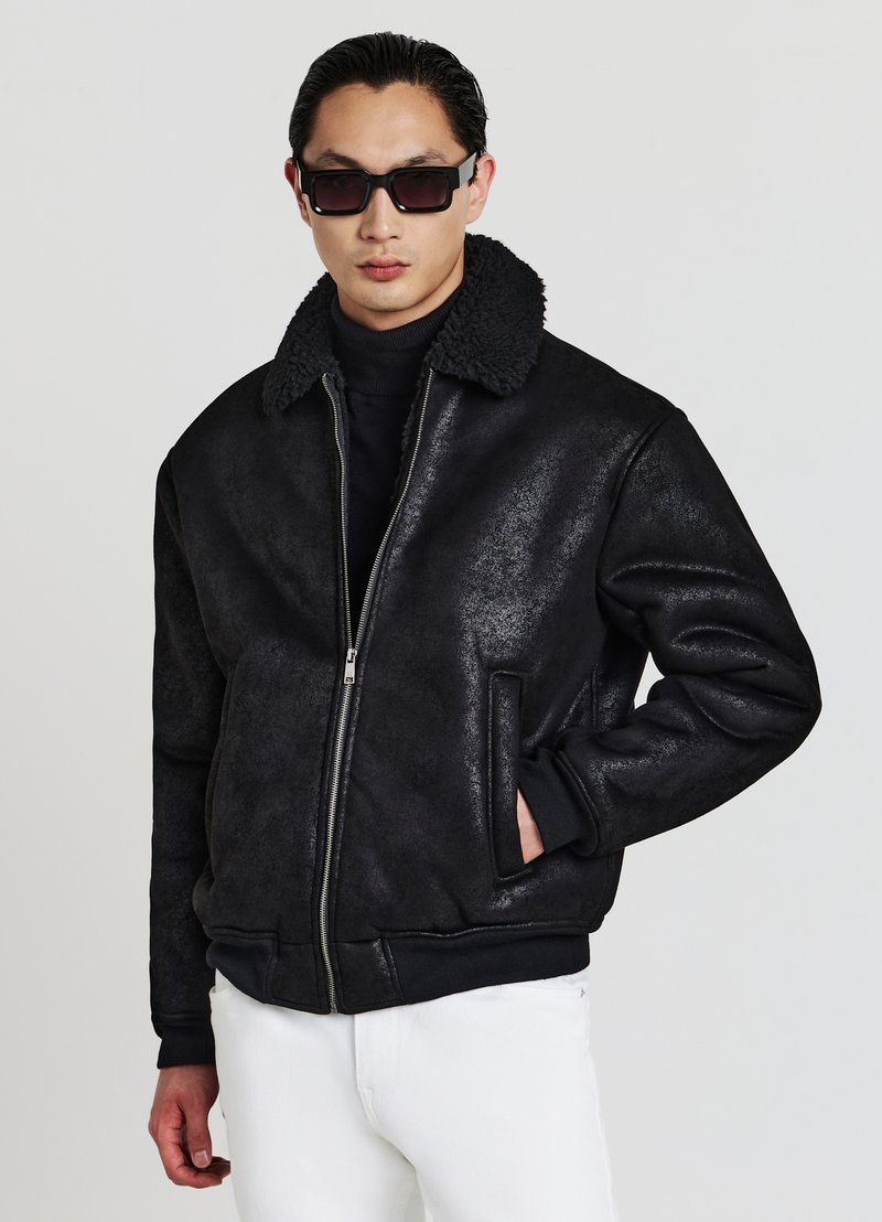 Schwarze Bomberjacke mit strukturiertem Finish und fellgefüttertem Kragen. Verfügt über einen Reißverschluss vorne und zwei seitliche Taschen. Trägt man über einem Rollkragenpullover.